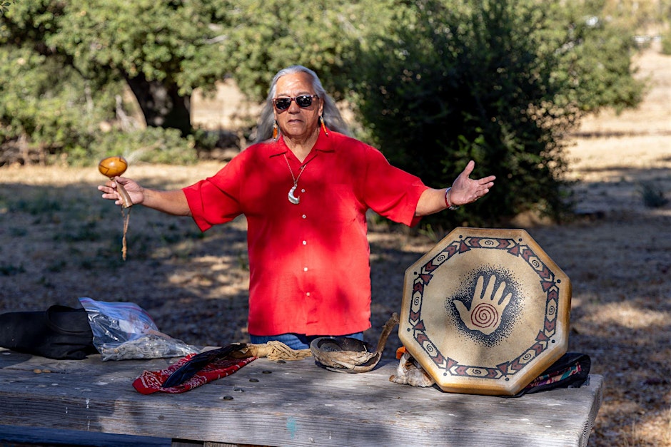 Chumash Elder Dennis Garcia standing outdoors behind a table with a drum and cultural items, leading a songs and stories workshop at Satwiwa.
