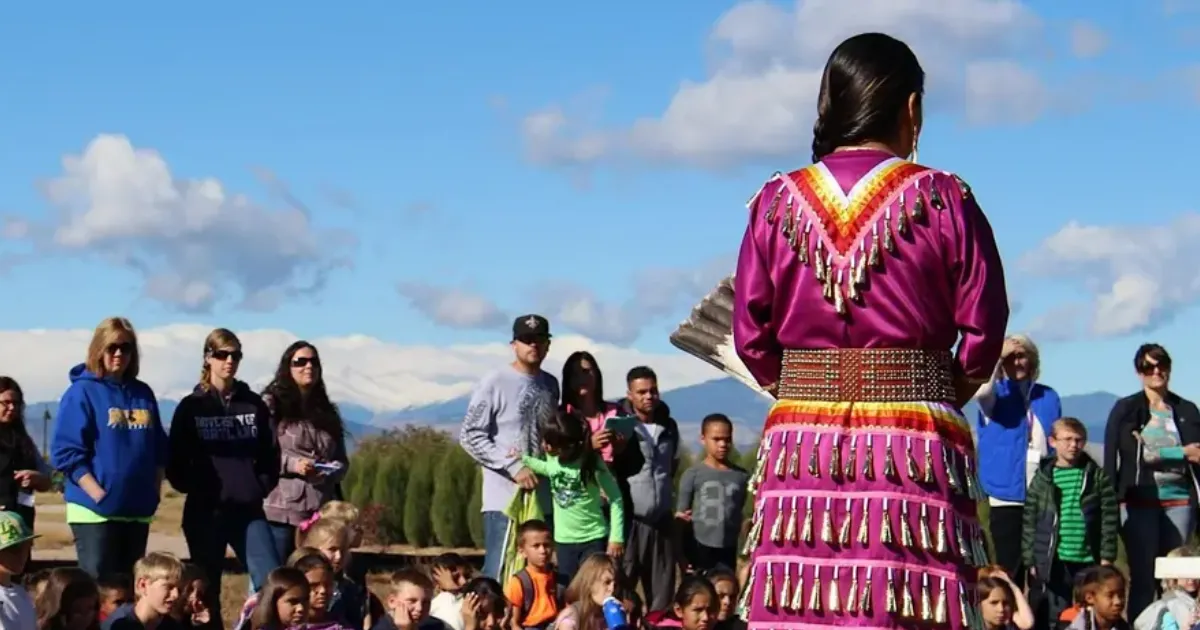 Native dancer in vibrant purple regalia presenting outdoors to families and community members during a Native American cultural program.